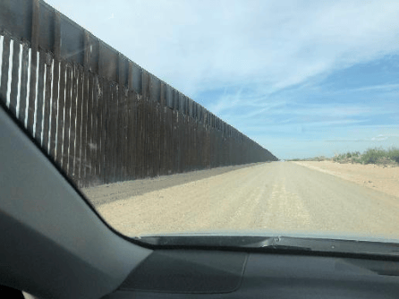 View from a vehicle along a dusty road beside a tall metal fence in a sparse desert landscape.