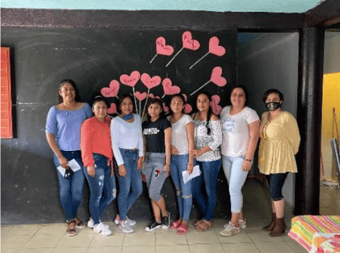Eight women pose before a blackboard decorated with pink hearts.