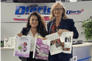 Two women hold magazines at a promotional counter beneath a blue-and-red logo.