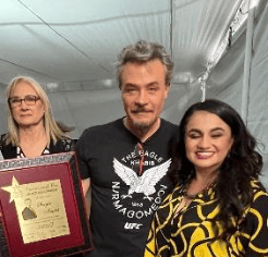 Three adults pose indoors, with one woman holding a framed plaque.