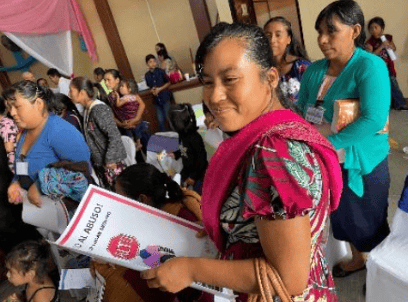 Smiling woman holding a white booklet in a crowded indoor gathering.