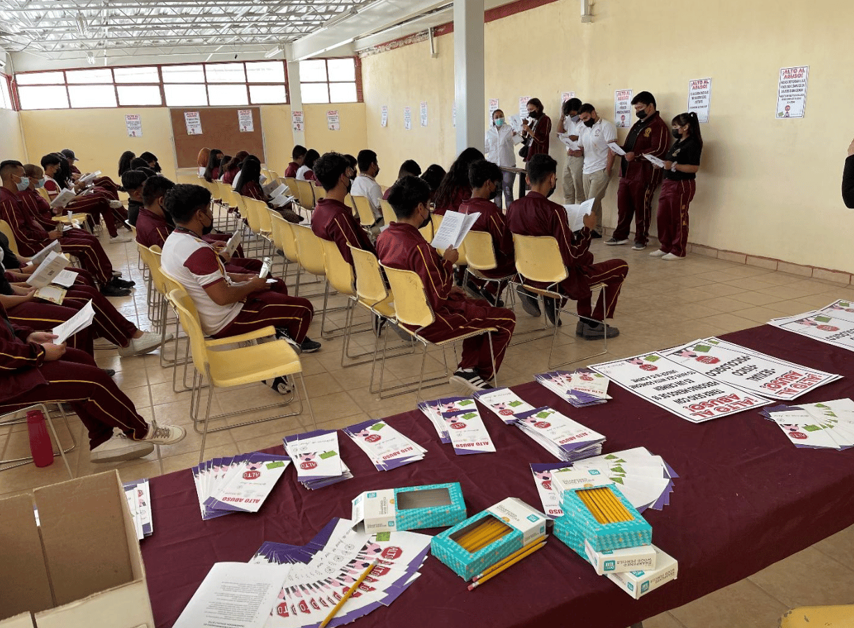 Masked attendees in maroon uniforms read handouts while presenters stand beside an outreach table.
