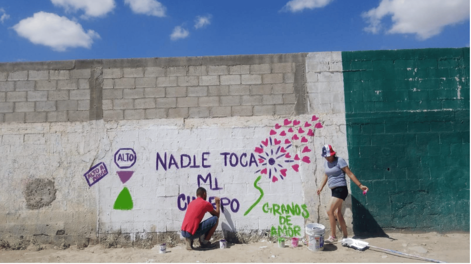 Two people paint a colorful Spanish message and flower mural on a cinder-block wall.
